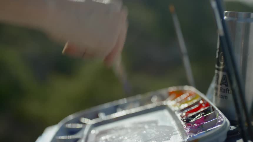 A woman wearing a hat and sunglasses painting onto a pad held up by an easel atop a mountain