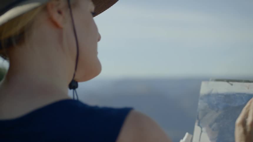 A female is blotting paint to blend it on a canvas set up on a painting easel set up outdoors with a view of the mountains and canyon in front of her