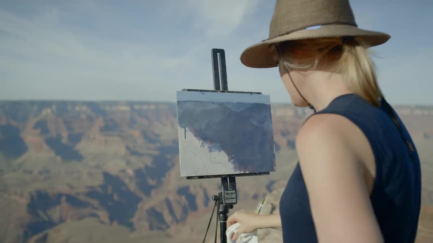 A white female artist is blending paint on a canvas held by a painting easel outside overlooking a canyon and mountains