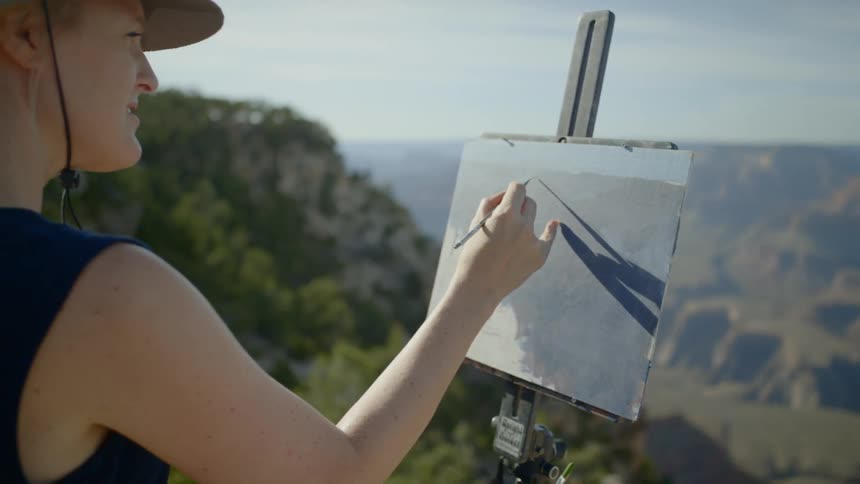 A woman is outdoors painting a landscape of a canyon and plateau