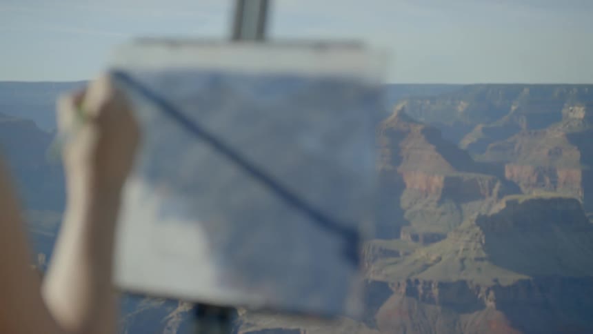 A woman wearing a hat and sunglasses painting onto a pad held up by an easel atop a mountain