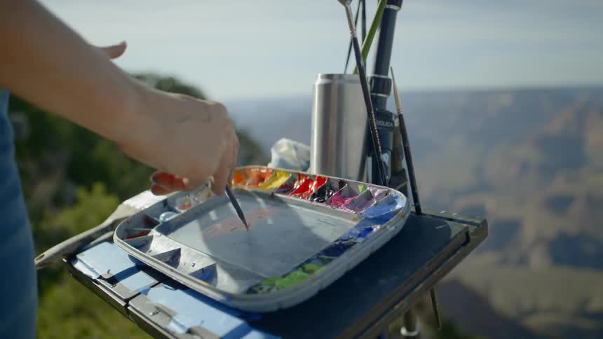 A woman standing next to a canyon mixing paint with a paintbrush