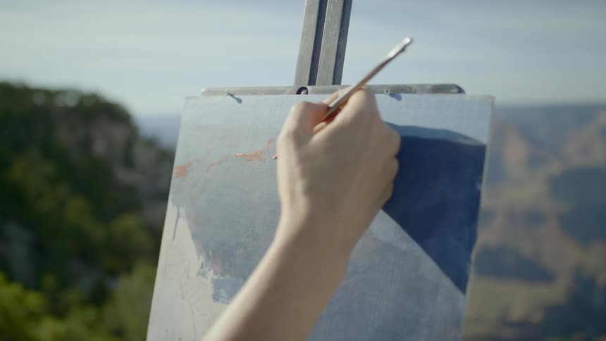 A woman wearing a hat is painting onto a canvas held up by an easel atop a mountain