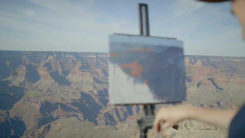 A woman wearing a hat is painting onto a canvas held up by an easel atop a mountain
