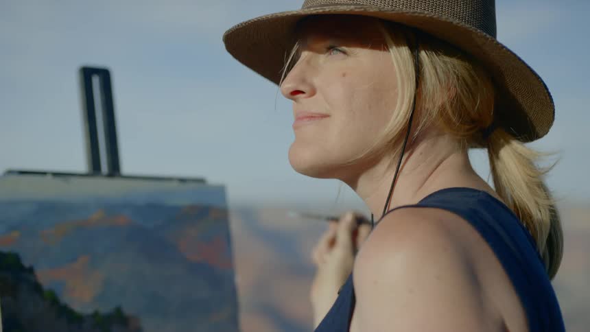 A woman stands outside and paints the mountains in front of her