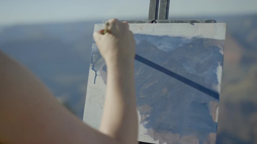 A woman wearing a hat and sunglasses paints onto a pad held up by an easel