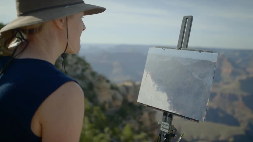 A blonde woman stands on the edge of a canyon and compares her view with a picture on a stand