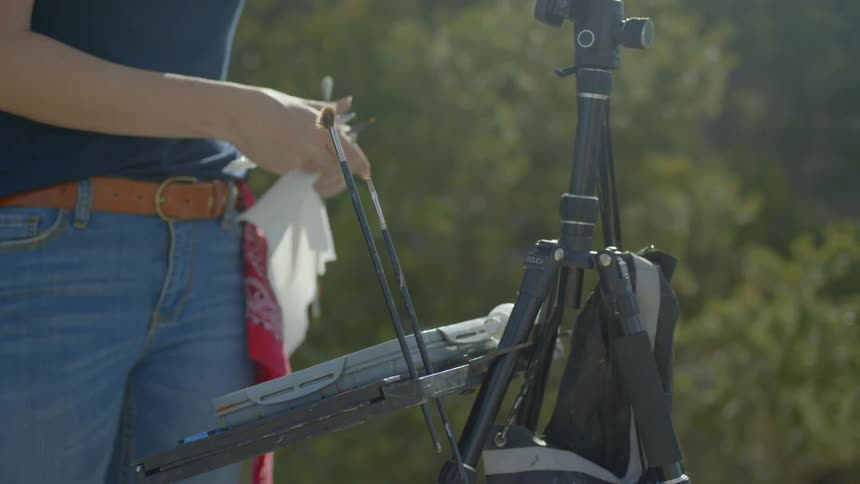 A woman places paintbrushes in holders on her outdoor easel