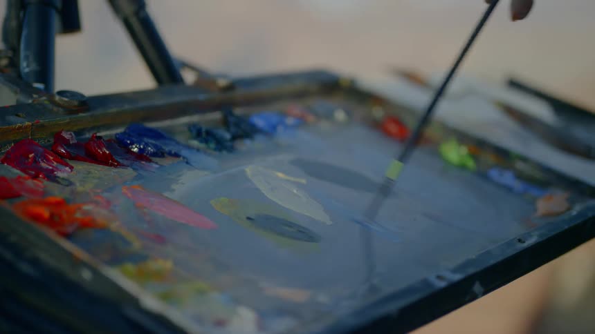 A woman wearing a hat is painting onto a canvas held up by an easel atop a mountain