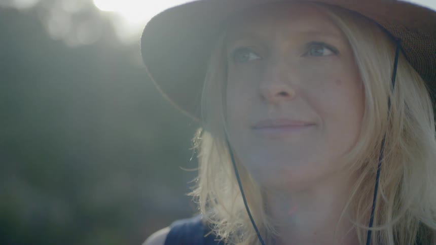 A blonde woman wearing a straw hat smiles as she looks around on a sunny day