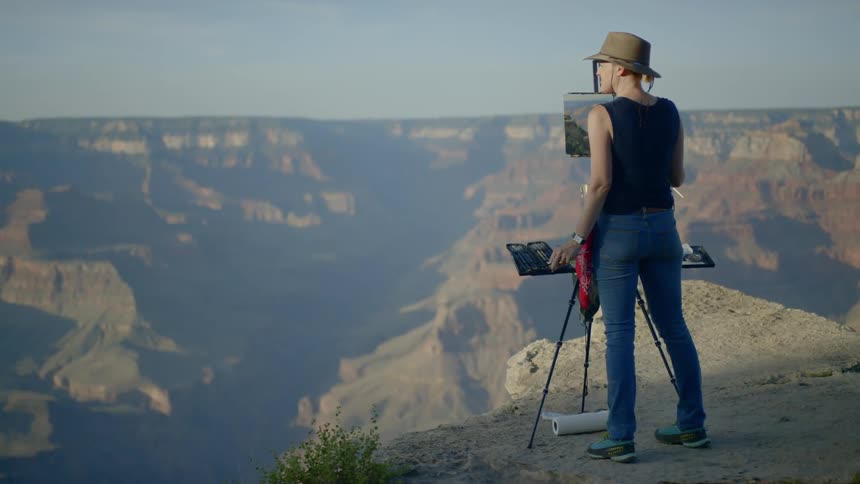 A woman wearing a hat is painting onto a canvas held up by an easel atop a mountain