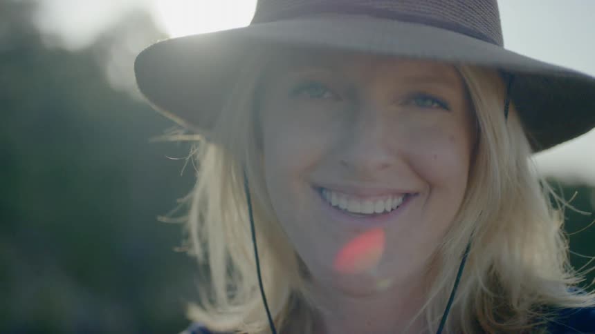 A blonde haired woman wearing a hat smiles as she looks directly into the camera while standing outside on a sunny day