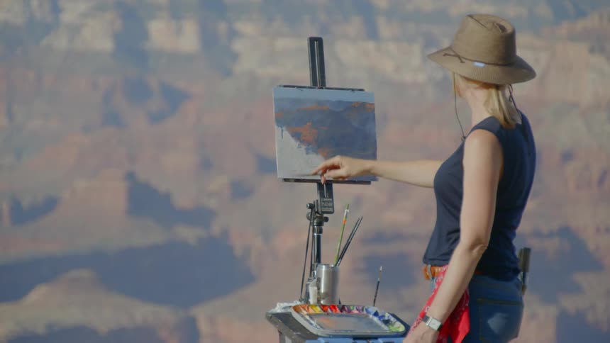 A woman wearing a hat is painting onto a canvas held up by an easel atop a mountain