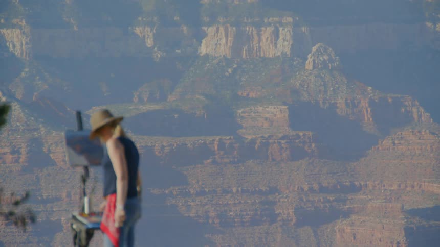 A white female is using a painting easel canvas and paints that are set up overlooking a view of mountains