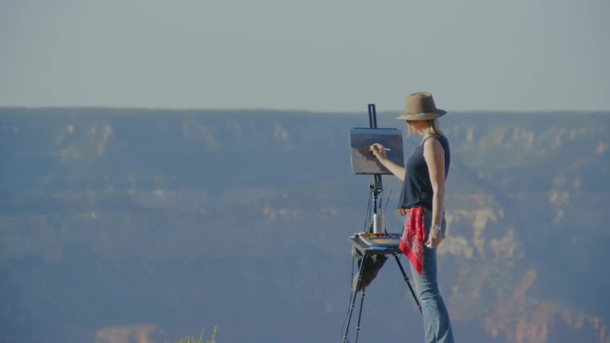 A woman wearing a hat is painting onto a canvas held up by an easel atop a mountain