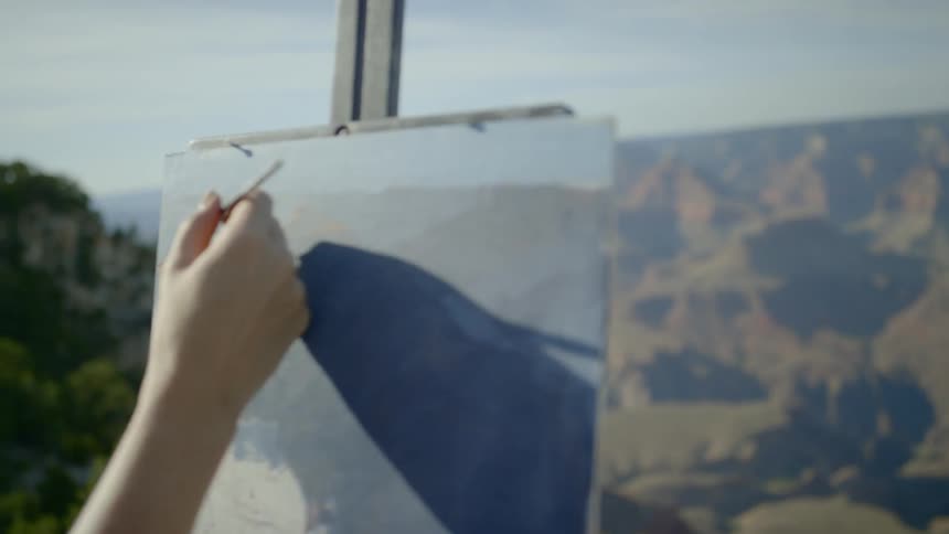An artist is using a fine point paint brush on a canvas on a painting easel to capture the view of the canyon and mountains before her
