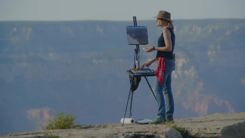 A woman wearing a hat is painting onto a canvas held up by an easel atop a mountain