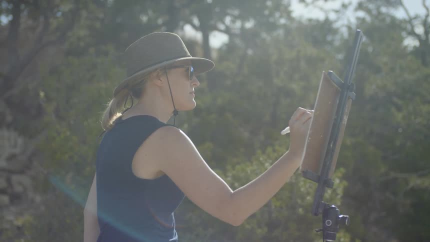 A woman wearing a hat and sunglasses sketches onto a pad held up by an easel