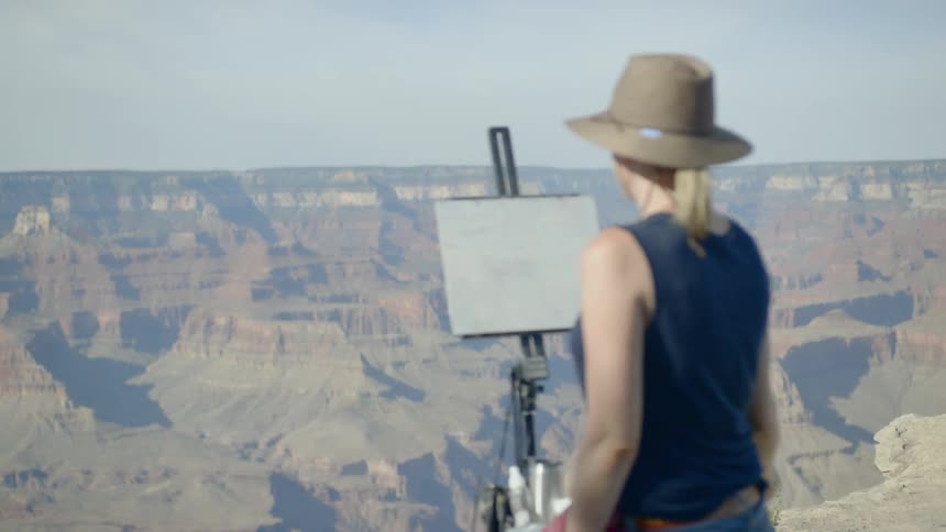 A woman wearing a hat and sunglasses painting onto a canvas held up by an easel atop a mountain