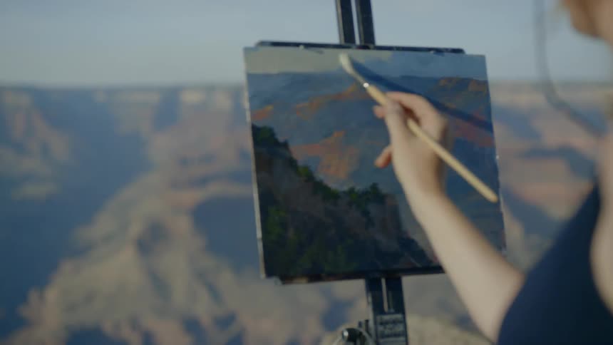 A woman wearing a hat is painting onto a canvas held up by an easel atop a mountain