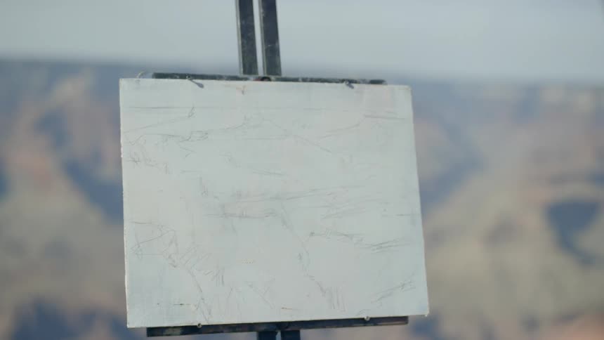 A woman wearing a hat is painting onto a canvas held up by an easel atop a mountain