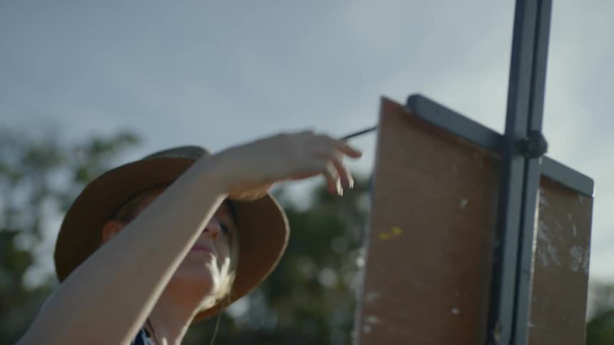 A woman wearing a hat is painting onto a canvas held up by an easel atop a mountain