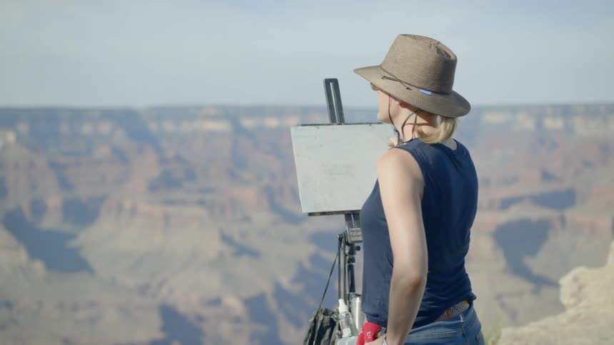 A woman wearing a hat and sunglasses painting onto a pad held up by an easel atop a mountain