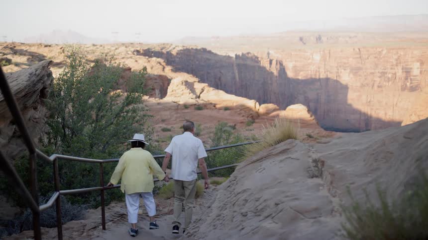 An old man and woman holding hands while walking along a path on the top of a canyon