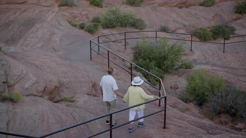 An older man and woman hold hands while walking down a rocky hill next to a handrail