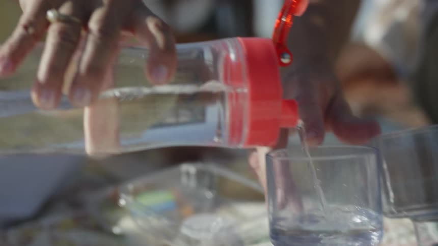 A woman sitting on a blanket outdoors pours water from a larger bottle into a small glass