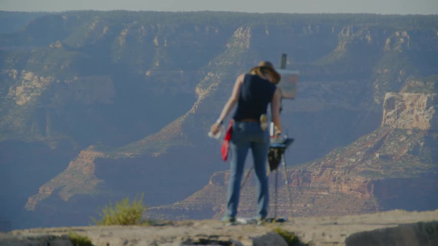 A woman wearing a hat is painting onto a canvas held up by an easel atop a mountain