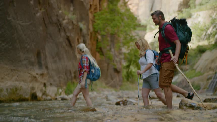 Three people in hiking gear and carrying backpacks are using walking sticks to cross a moving stream surrounded by mountains