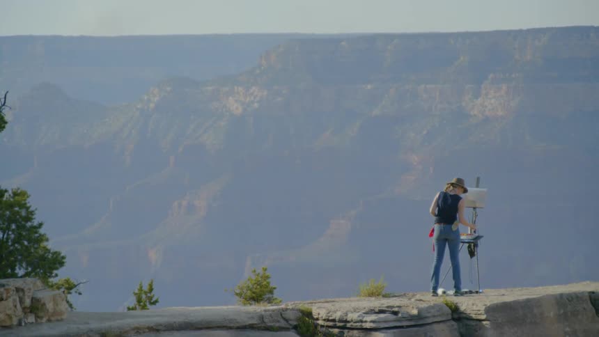 A woman wearing a hat is painting onto a canvas held up by an easel atop a mountain