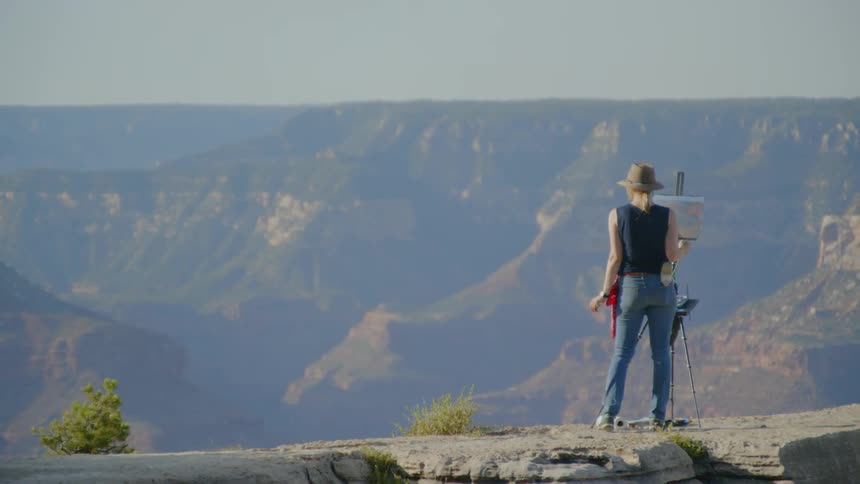 A woman paints a canvas standing on a mountain cliff