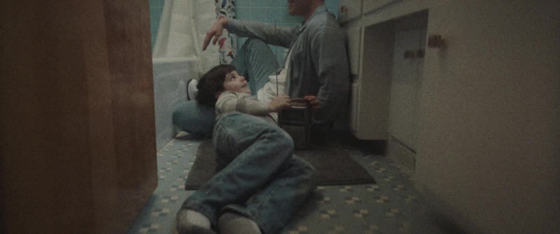 A young boy reclines on the bathroom floor with his head resting on his fathers leg as they listen to an older battery operated radio