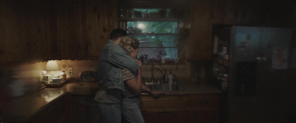 A young woman stands in a kitchen and a man comes up behind her and hugs her
