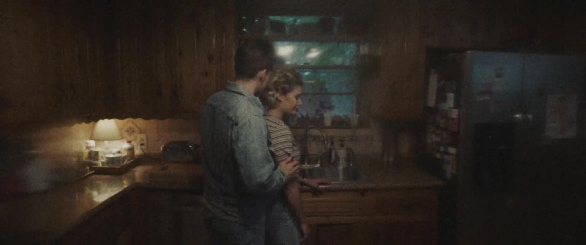 Blonde woman stands next to the sink and man approaches her to cuddle in the kitchen at night