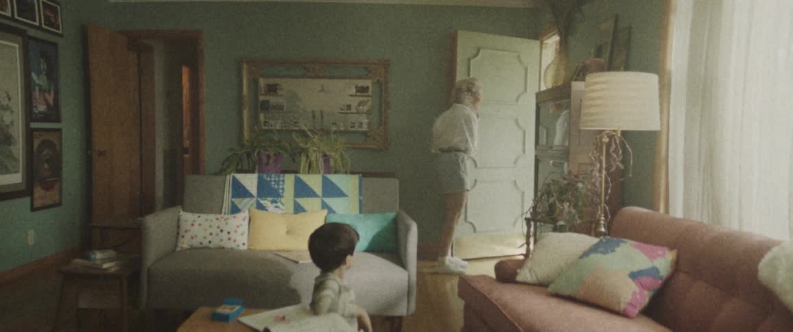 A woman is walking through a living room to answer the front door as a young boy sits on the floor doing homework and then a man enters the door