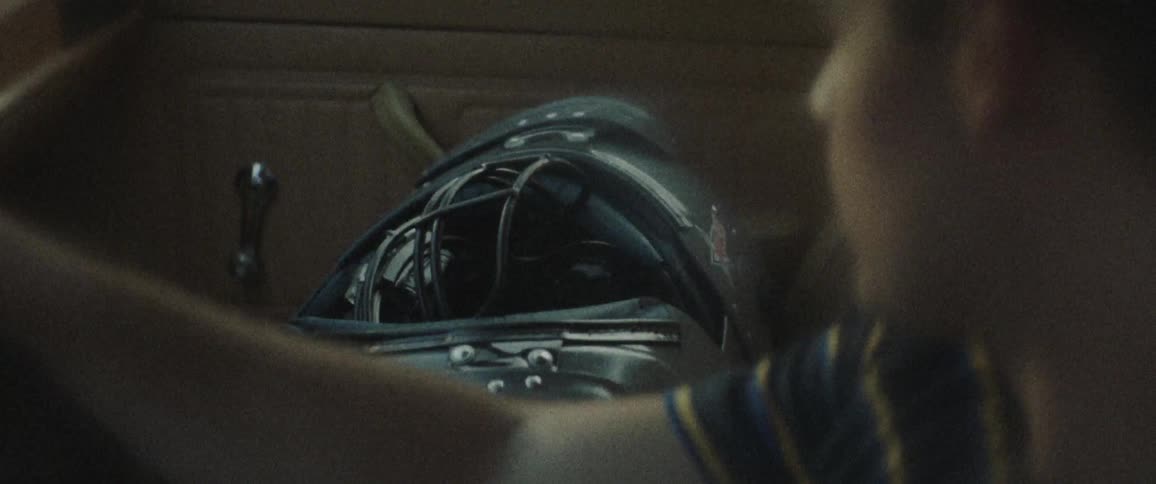 A young man driving a car looks over at the passenger seat where his shoulder pads and helmet are stacked