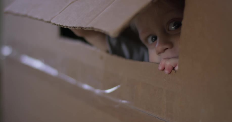 A young child smiles and peaks out of a cardboard box