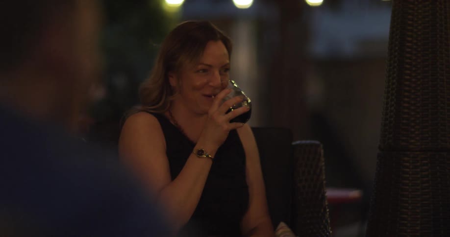 A woman sips from a wine glass while laughing next to a fire