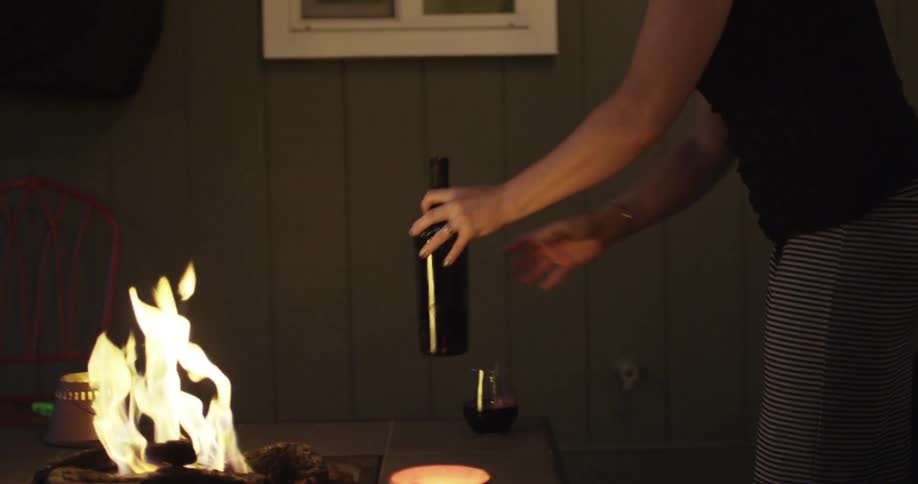 A woman pours a bottle into a glass by a fire