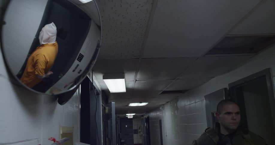 A soldier leads a hooded prisoner down a hall and past a reflective mirror