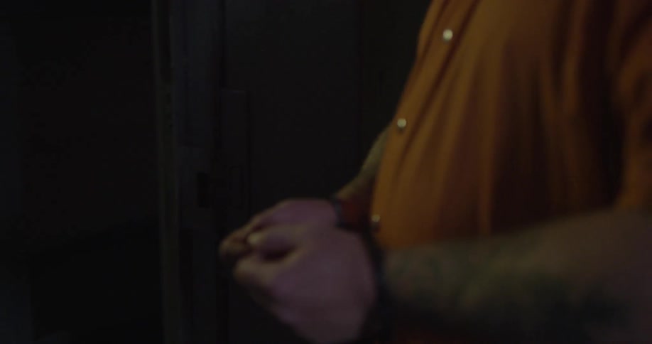 A handcuffed man in an orange suit is led into a cell