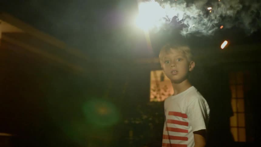 A blond haired child plays with a sparkler in front of a house at night