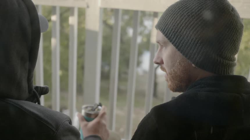 Two men in hats and jackets are talking on a balcony and one has a canned beverage in his hand