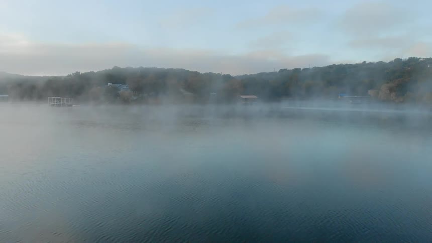 Fog hangs above the surface of a large body of water next to a forested shoreline