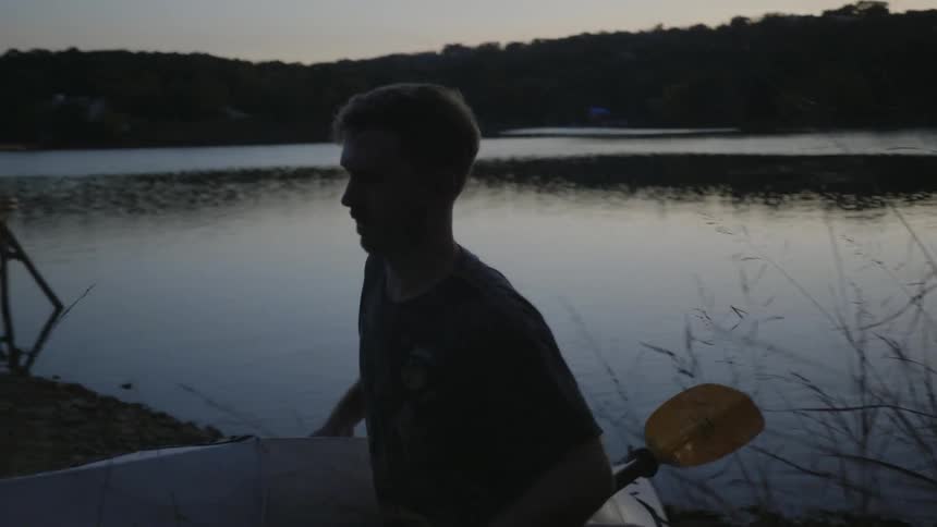 A white male is walking away from a calm lake carrying a canoe with his right hand and paddle with this left at dusk