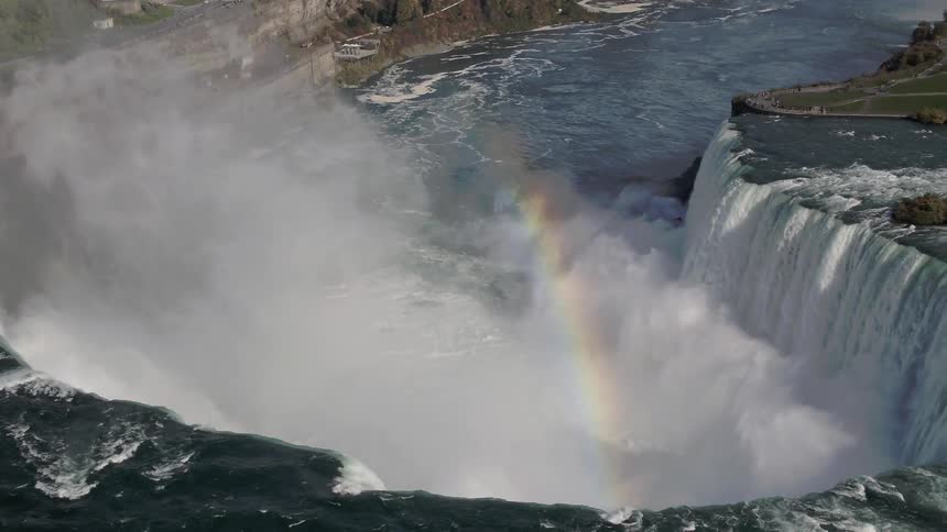 A rainbow forms over the water in front of a waterfall