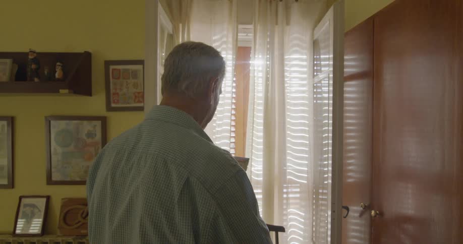 Sunshine beams through blinds and shines on a man standing in a room near a window